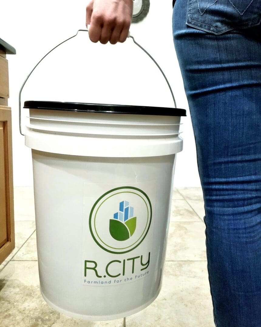A person in jeans holds a white bucket by the handle. The bucket features the R. City logo and the slogan Farmland for the Future. The scene appears to be indoors on a tiled floor.