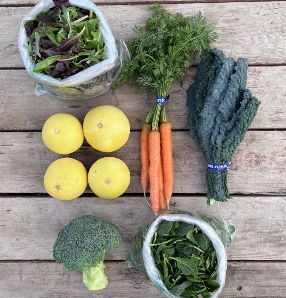 An assortment of fresh produce on a wooden surface: a bag of leafy greens, three yellow grapefruits, a bunch of carrots with tops, two bunches of dark kale, a head of broccoli, and a bag of spinach.