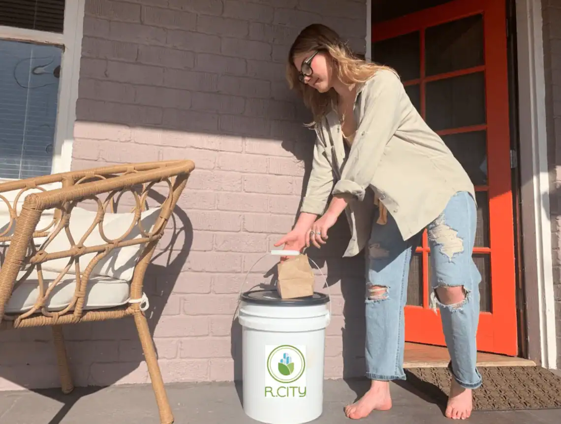 A woman in ripped jeans and a beige shirt places a brown paper bag into a white R.City compost bin on a porch next to a wicker chair and an orange door.